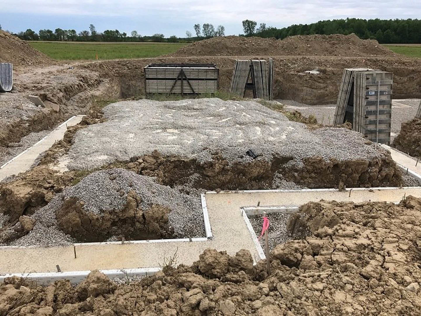 Gravel piles and wooden beams scattered across dirt foundation at residential construction site, with tree and sky visible in background