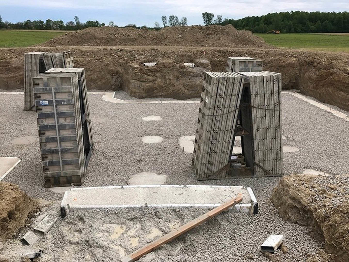Construction site with stacked wooden crates and planks on dirt ground, scattered rocks, concrete surfaces, and surrounding trees under a cloudy sky