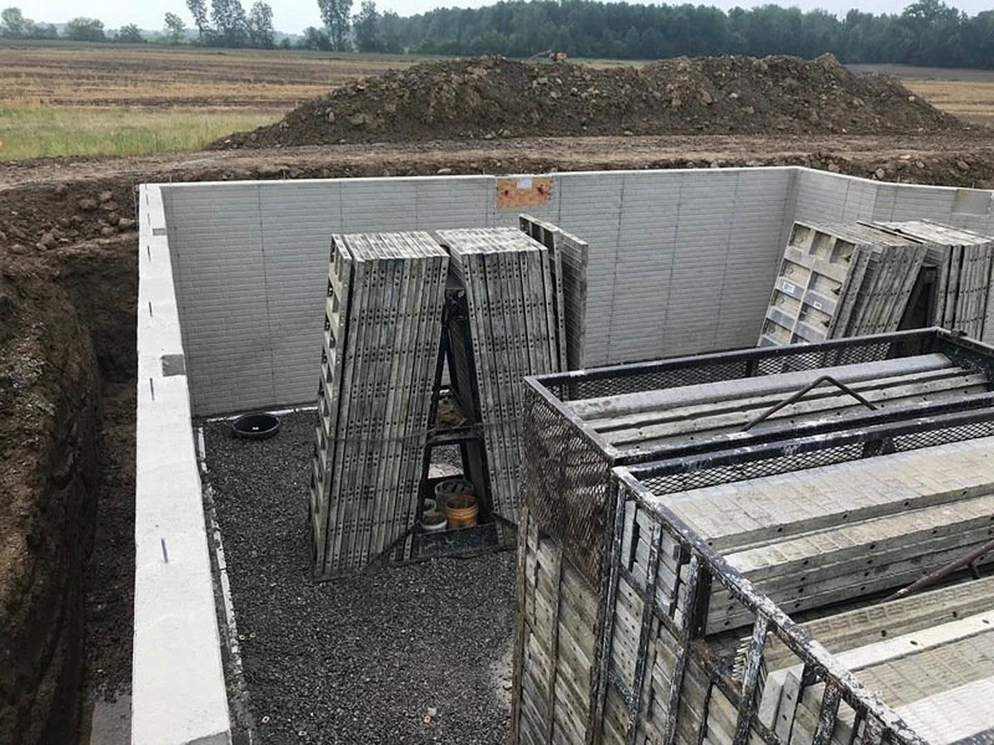 Concrete piles, metal scaffolding, and containers scattered across a construction site with dirt, rocks, and wood debris; fenced perimeter, trees, and grass visible in the