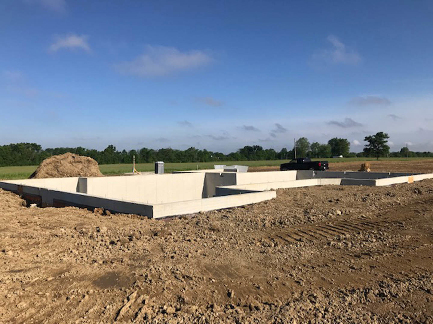 Concrete foundation set in a dirt lot surrounded by trees, under a blue sky with scattered clouds