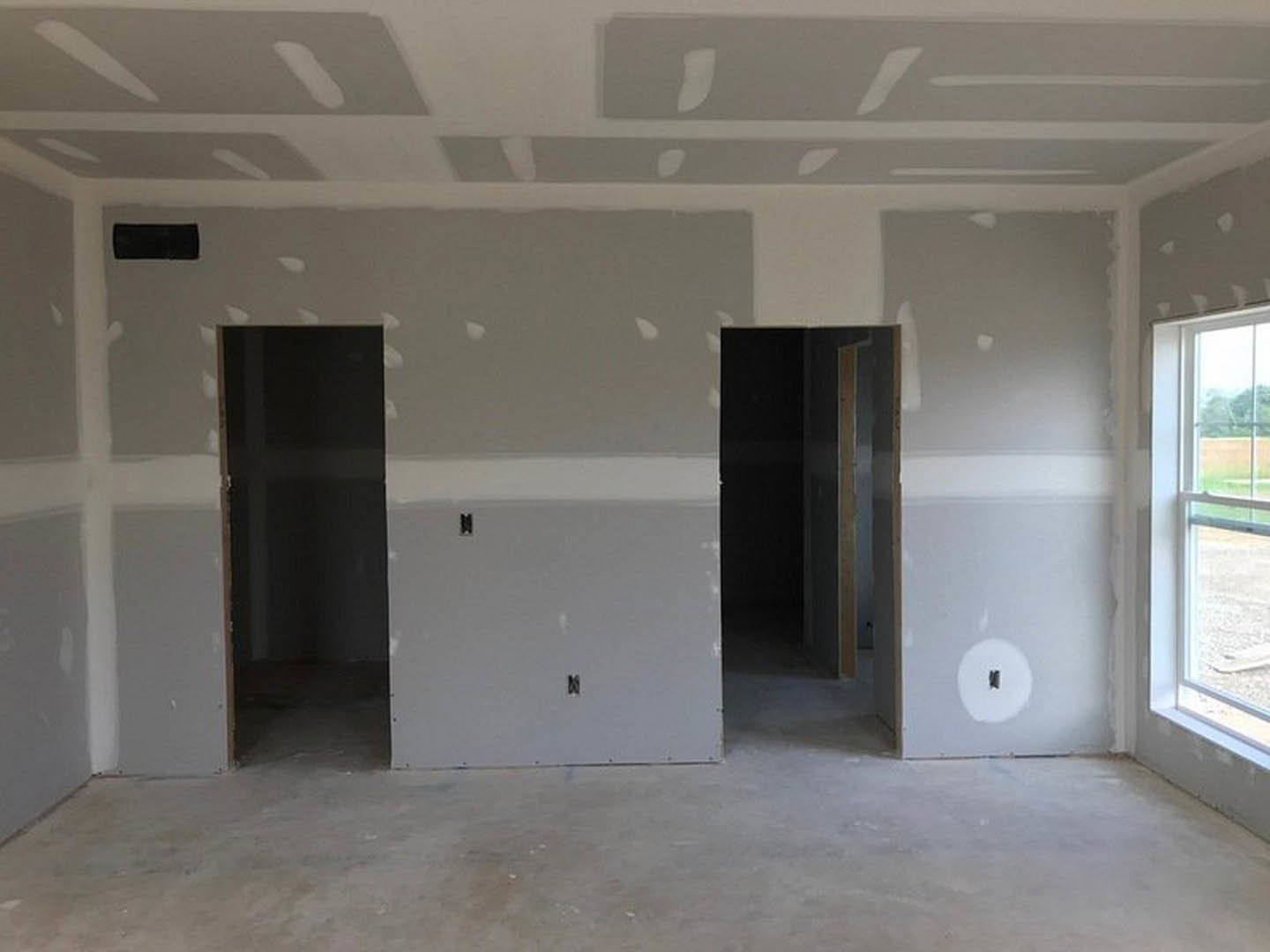 Bright room with smooth white walls, white doors, light flooring, and a window framed in white trim; black rectangular object and white board with black tape visible near door.