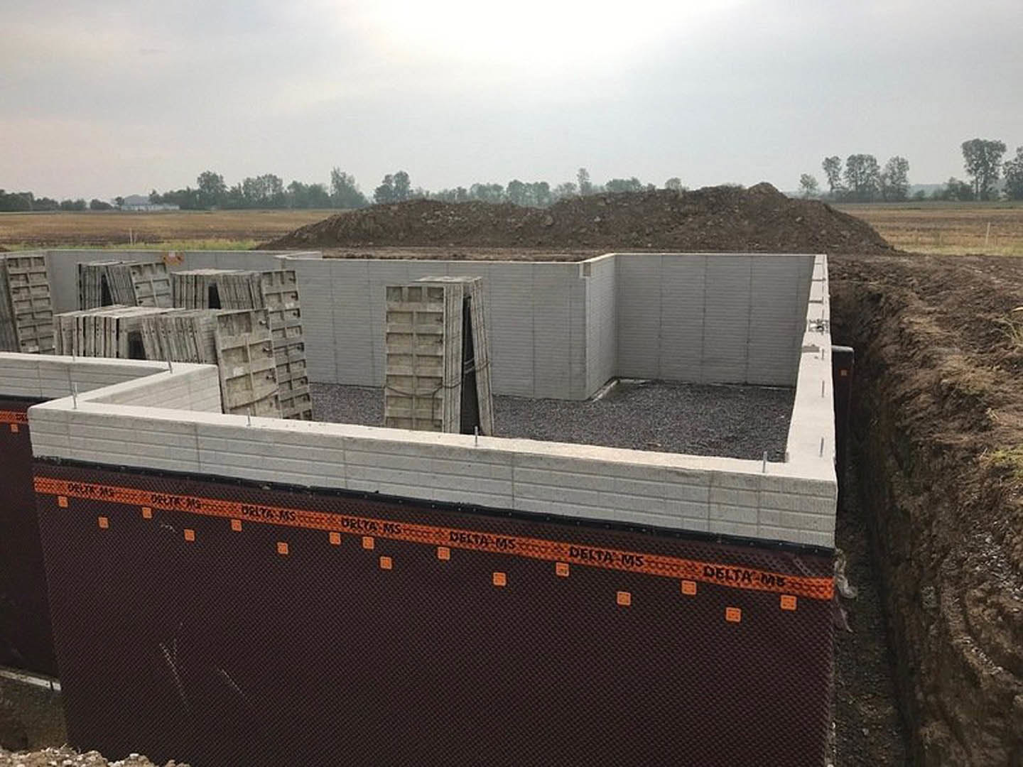 Framed custom home under construction with exposed concrete walls, dirt foreground, and trees in the background beneath a cloudy sky