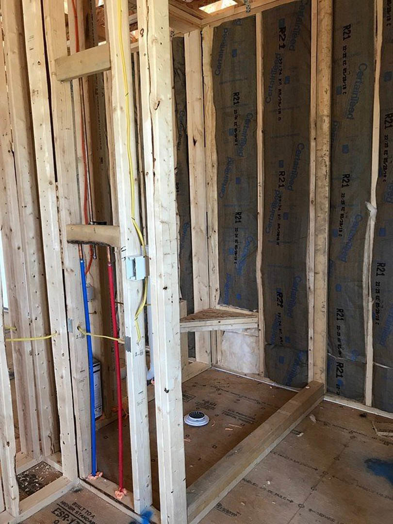 Exposed wood framing with blue and red plumbing pipes, electrical box, unfinished subfloor with drain, and partially insulated walls in a room under construction