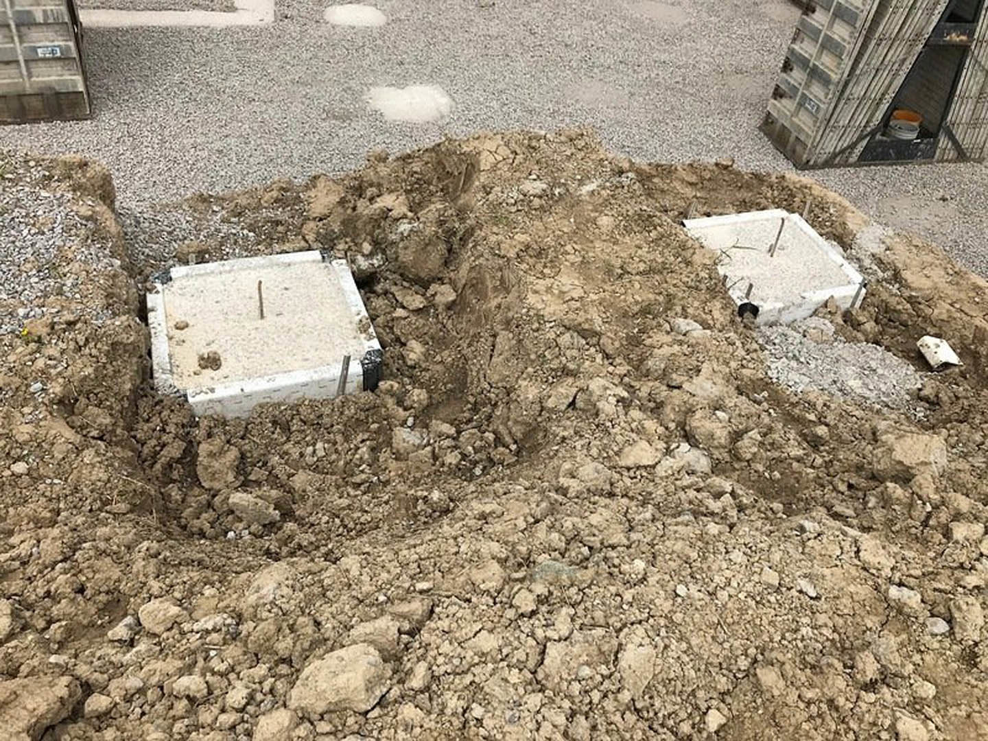 Pile of loose soil with two square concrete blocks, one embedded with a metal rod, at an outdoor construction site