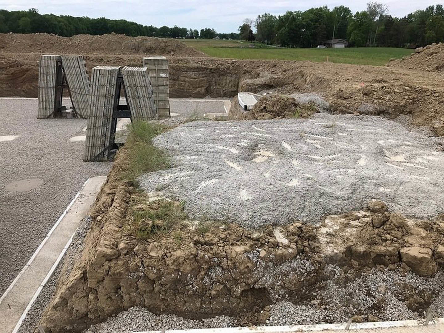 Cement and concrete pile on dirt and grass, concrete drainage channel on rocky ground, scattered rocks and soil, house in background, distant trees, white boat resting on ground