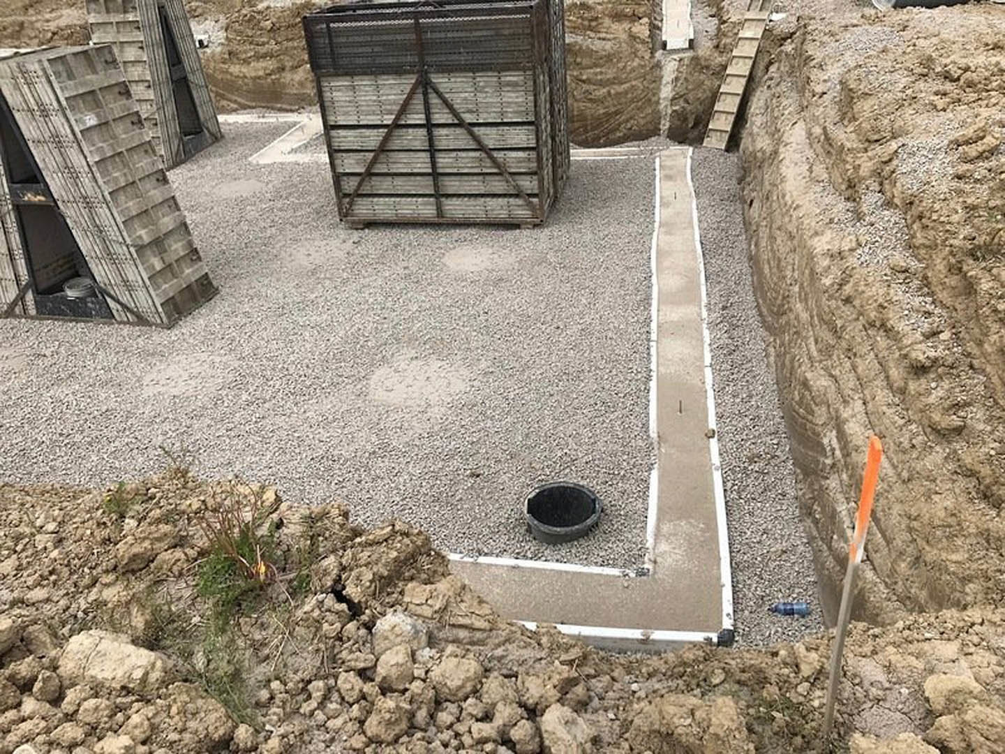 Excavated construction site with exposed soil and rocks, wooden formwork with metal reinforcement, black pipe on ground, concrete foundation in progress