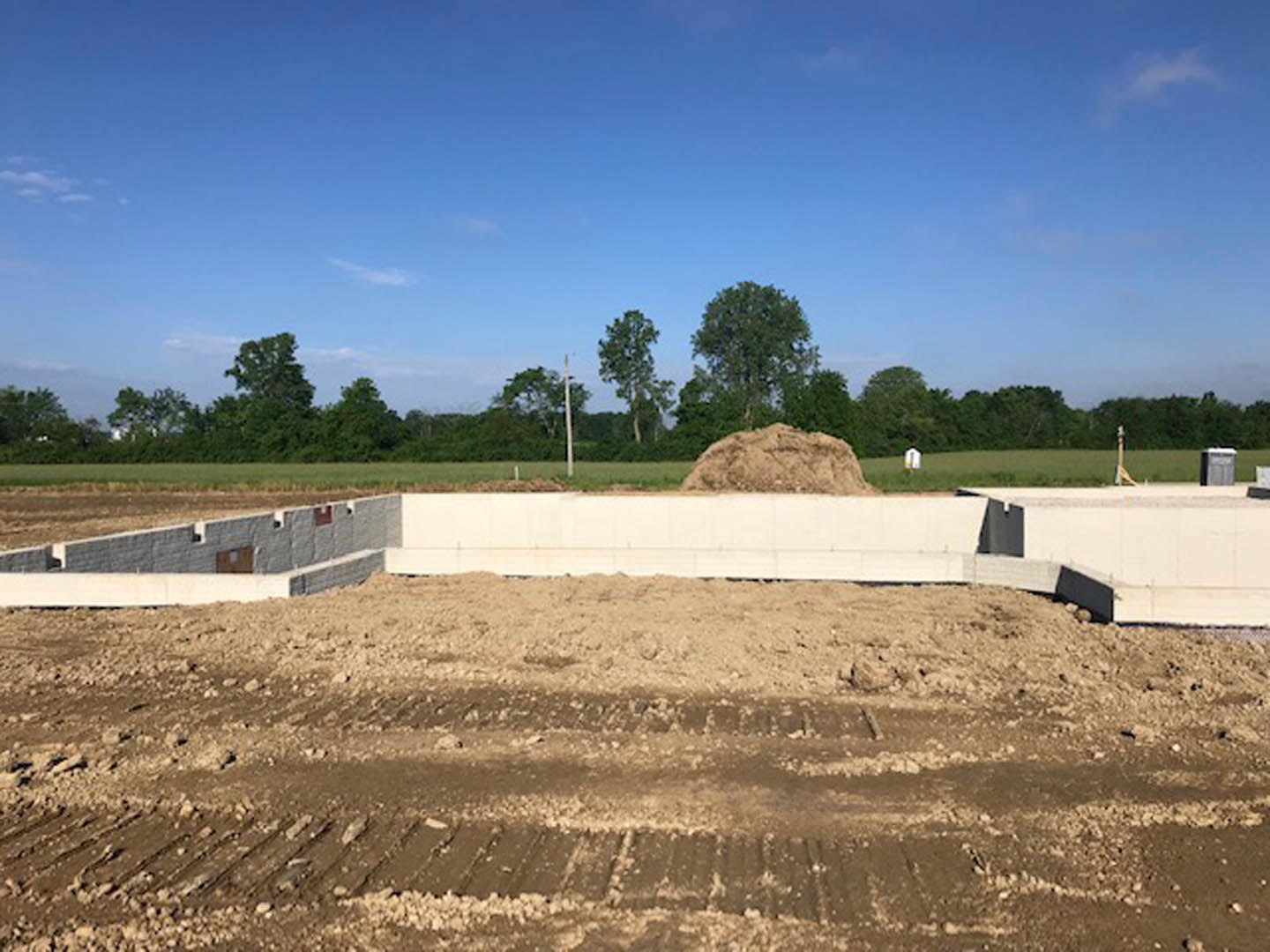 Large dirt pile with tire tracks in an open field, person standing nearby, green grass and group of trees in the background, blue sky with scattered clouds overhead
