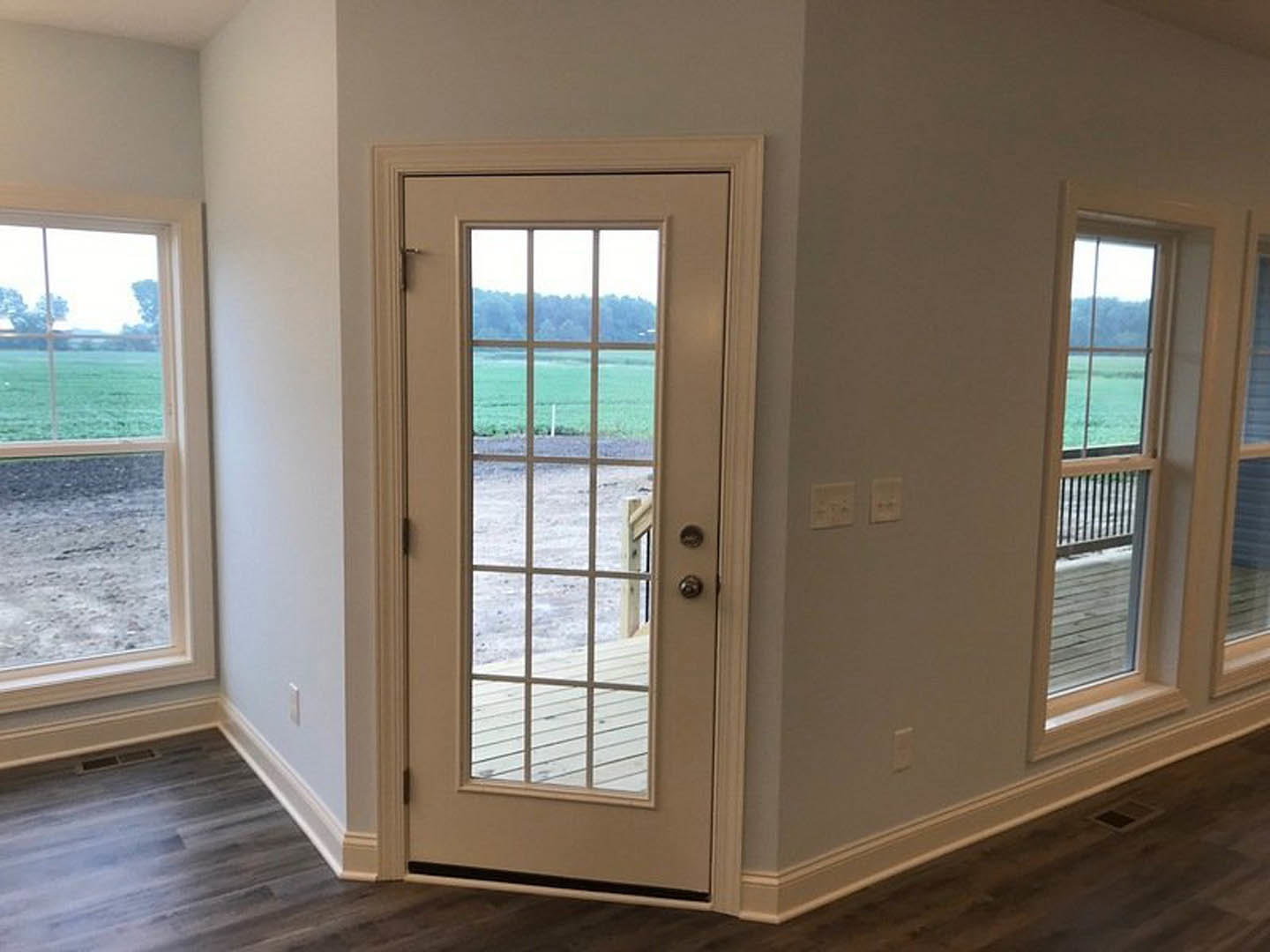 Wooden door with silver handle in a bright room, large windows overlooking grassy field, white wall switch, light wood flooring