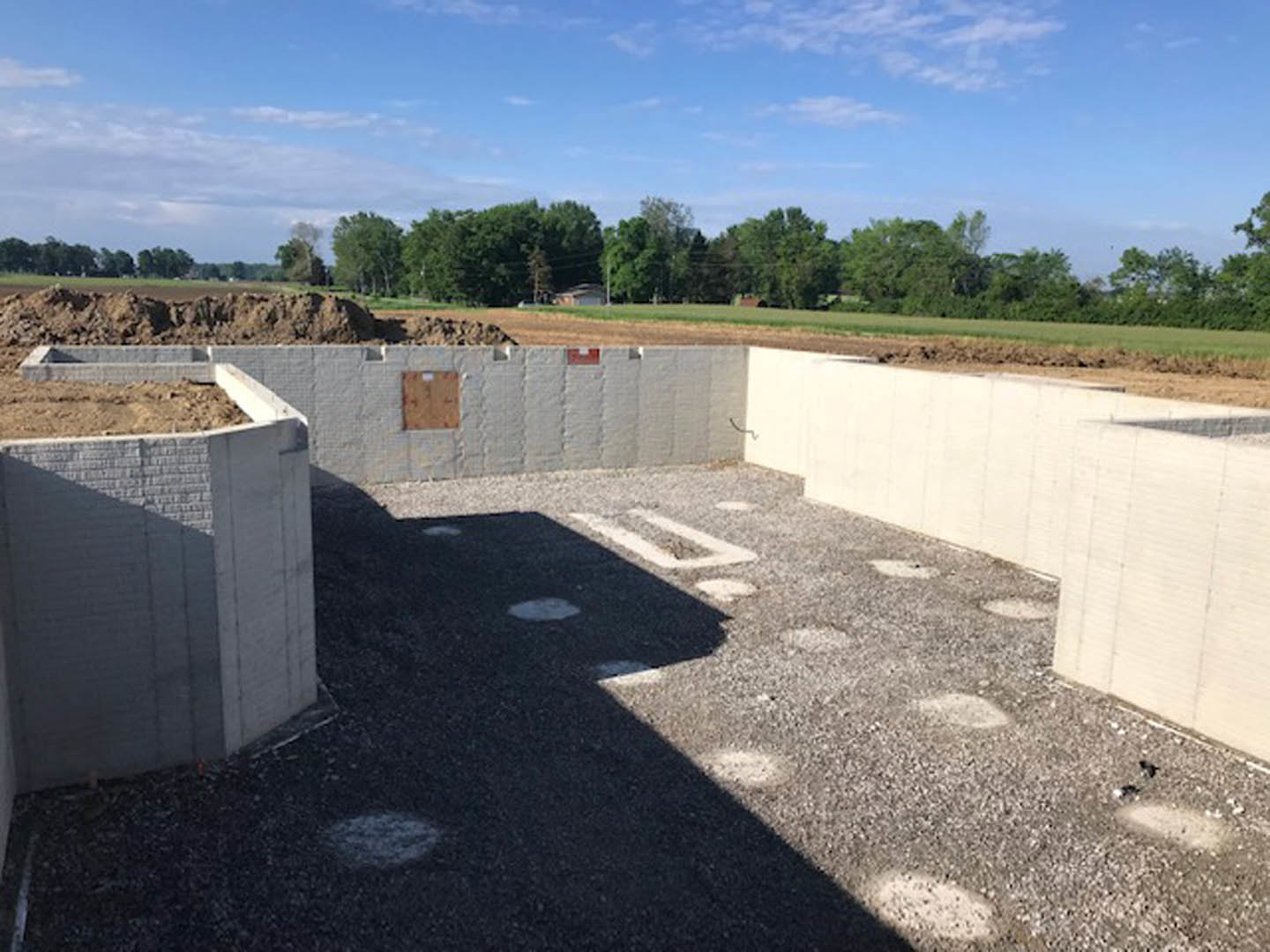 Concrete foundation slab surrounded by dirt, circular markings on surface, brown utility box resting on white concrete, blue sky with scattered clouds above green trees in
