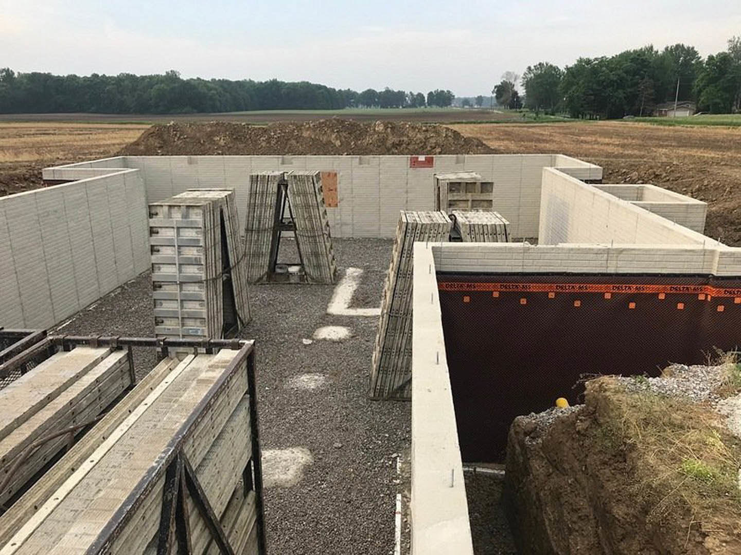 Wooden pallets stacked on gravel foundation, metal ladder leaning against framed structure, unfinished exterior surrounded by grass and rocks under partly cloudy blue sky