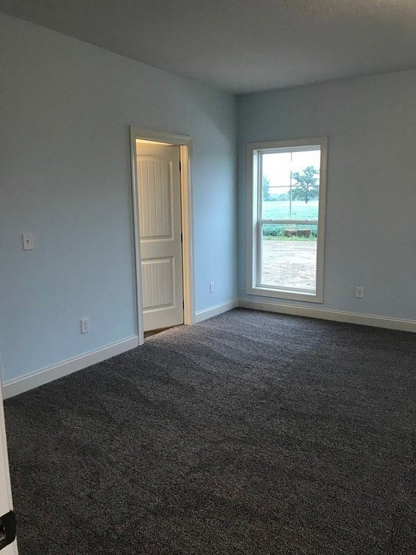 Carpeted room with white paneled walls, open white door, large window overlooking green field and leafy tree