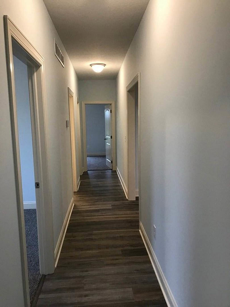 Hallway with medium-toned wood flooring, white walls, recessed ceiling light, and a white door with a black handle
