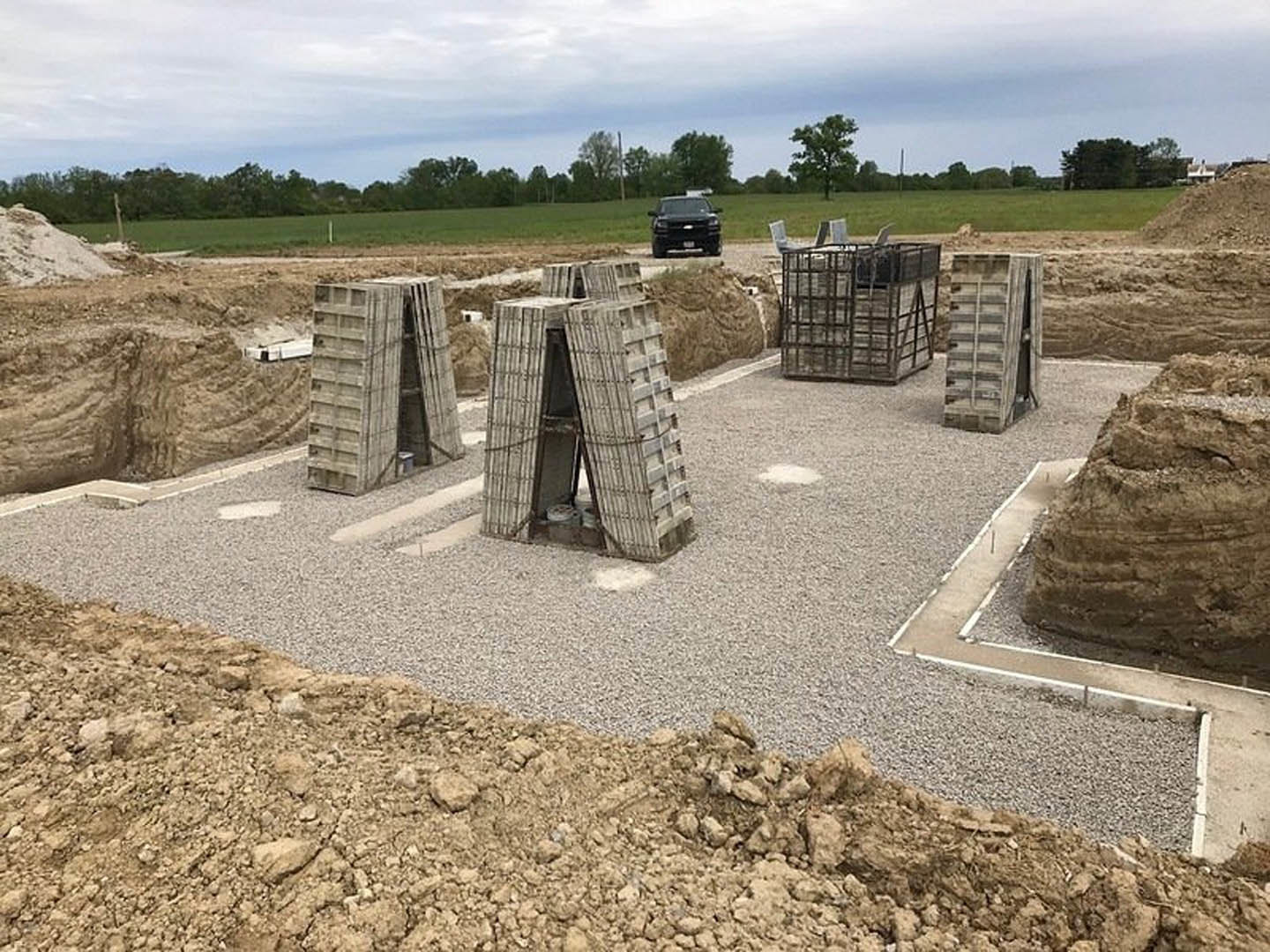 Concrete block foundation under construction, black car parked on dirt road, metal cage with chairs, stacked concrete slabs, trees and grass in background, cloudy sky overhead