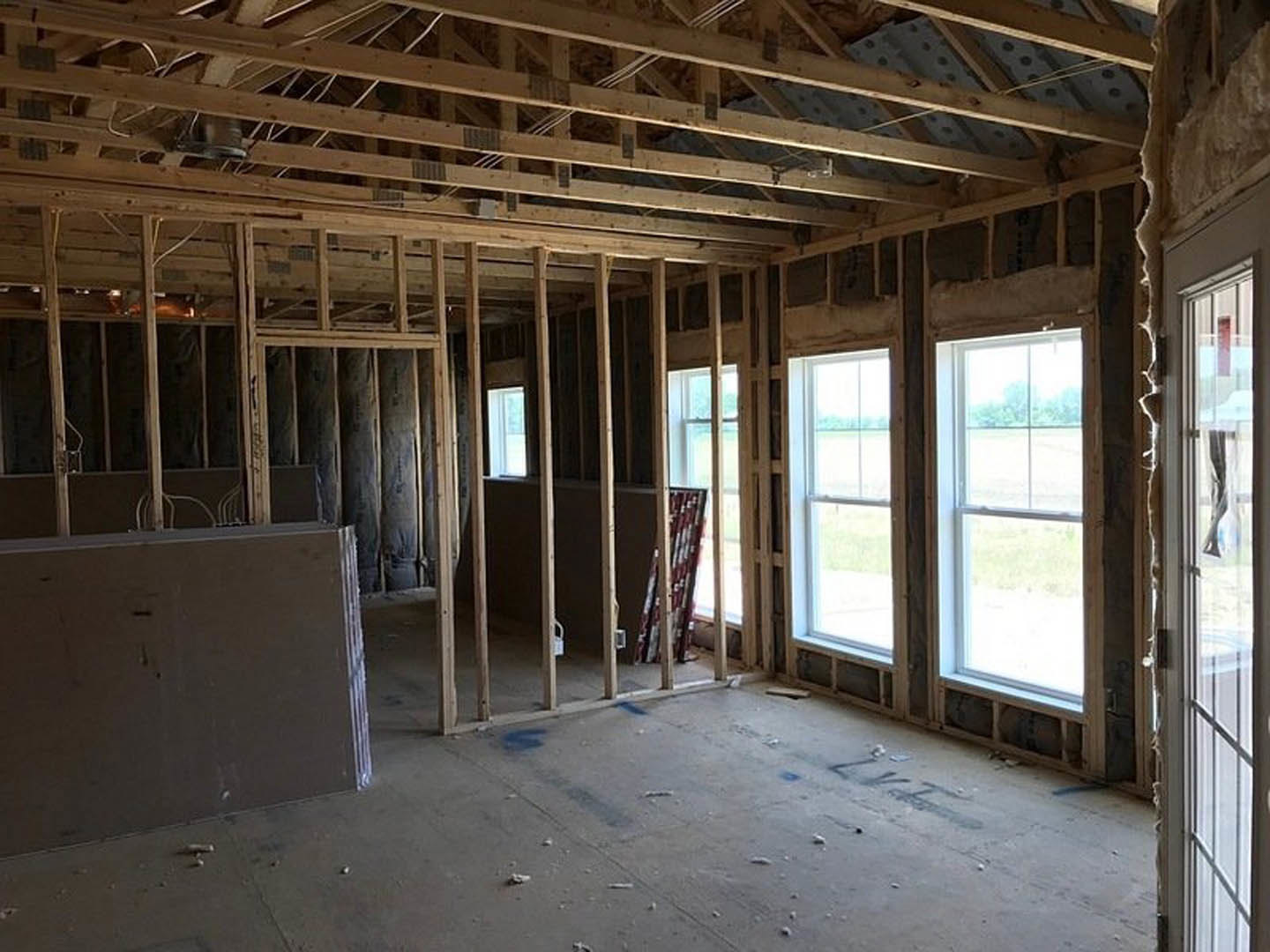 Open room with exposed wood ceiling beams, large windows overlooking green field, concrete floor with blue paint, unfinished wall with visible opening.