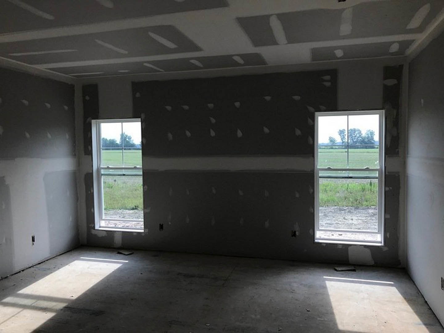 Neutral-toned room with two large windows, sunlight illuminating hardwood floor, white walls, and view of green grass and trees outside