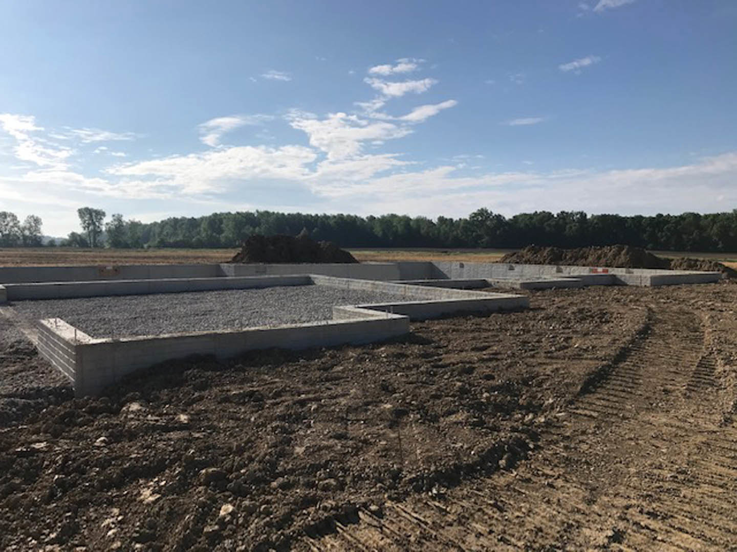 Concrete foundation set on a dirt construction site, surrounded by trees and soil under a blue sky with scattered clouds