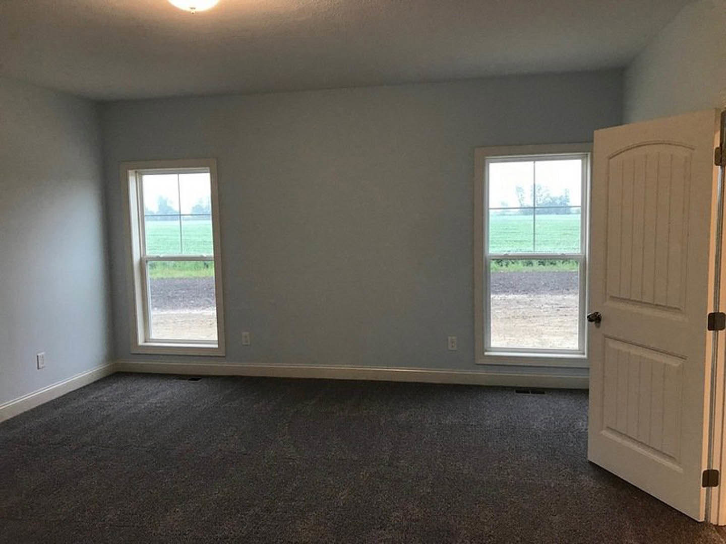 White plaster walls and light carpet flooring in a room with two windows overlooking a grassy field, open wood door visible on the left.