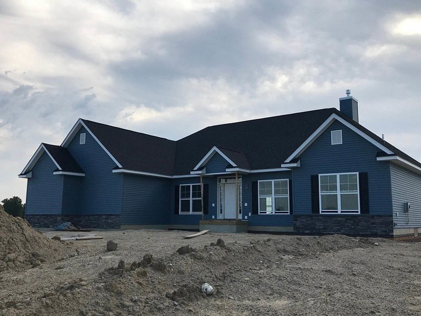 Partially built house with blue roof, white-framed window, exposed dirt foundation, scattered rocks, and cloudy sky overhead