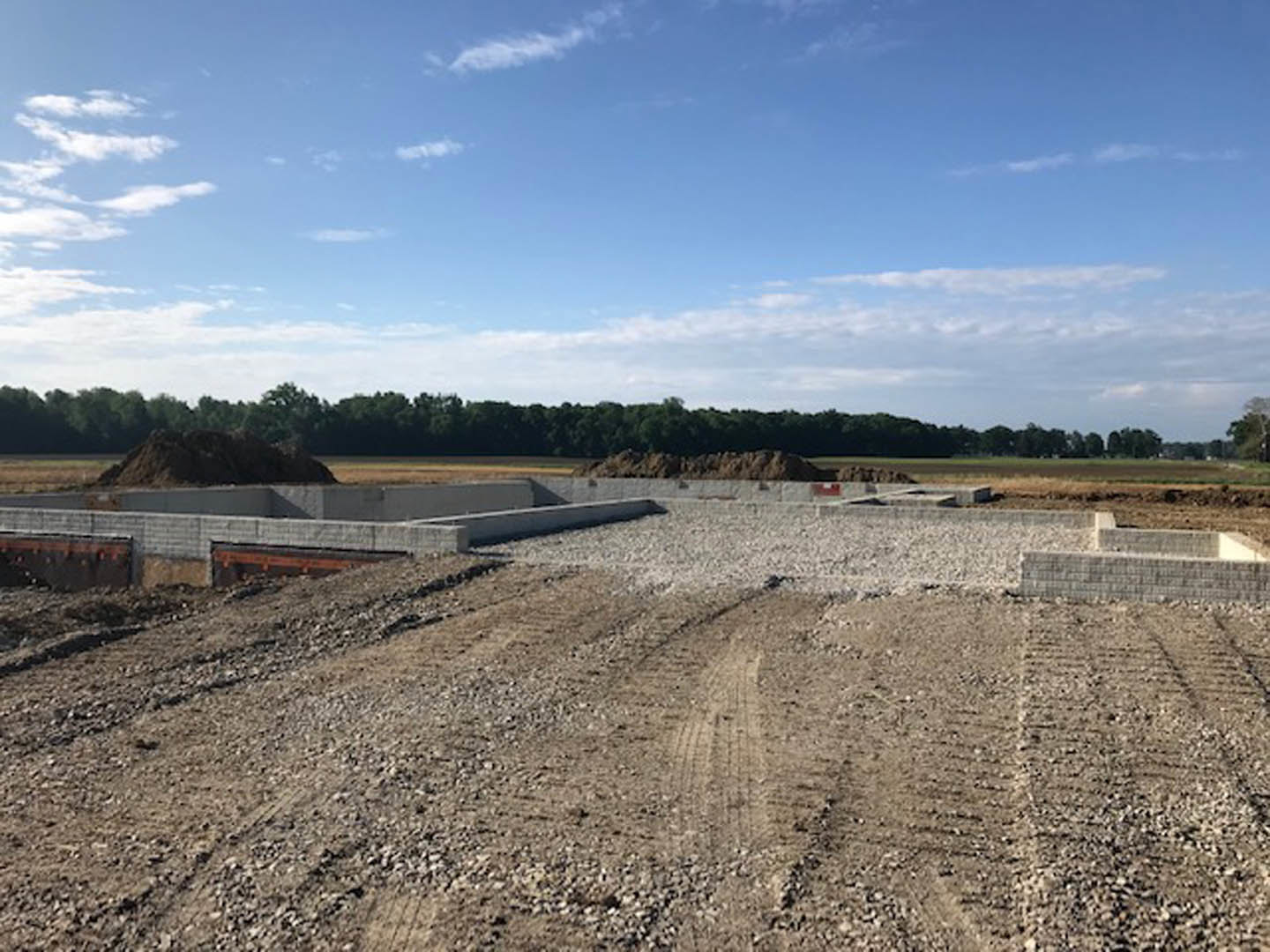 Dirt road with tire tracks leading through a construction site, bordered by soil and scattered trees under a blue sky with clouds; distant trees and a blurry black animal visible