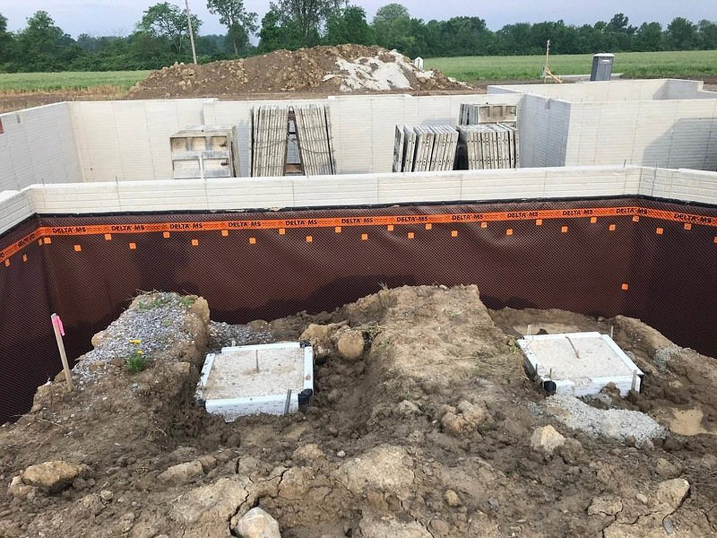 Concrete foundation surrounded by dirt, wooden stakes, and a metal fence, with scattered rocks and construction materials on an open field.