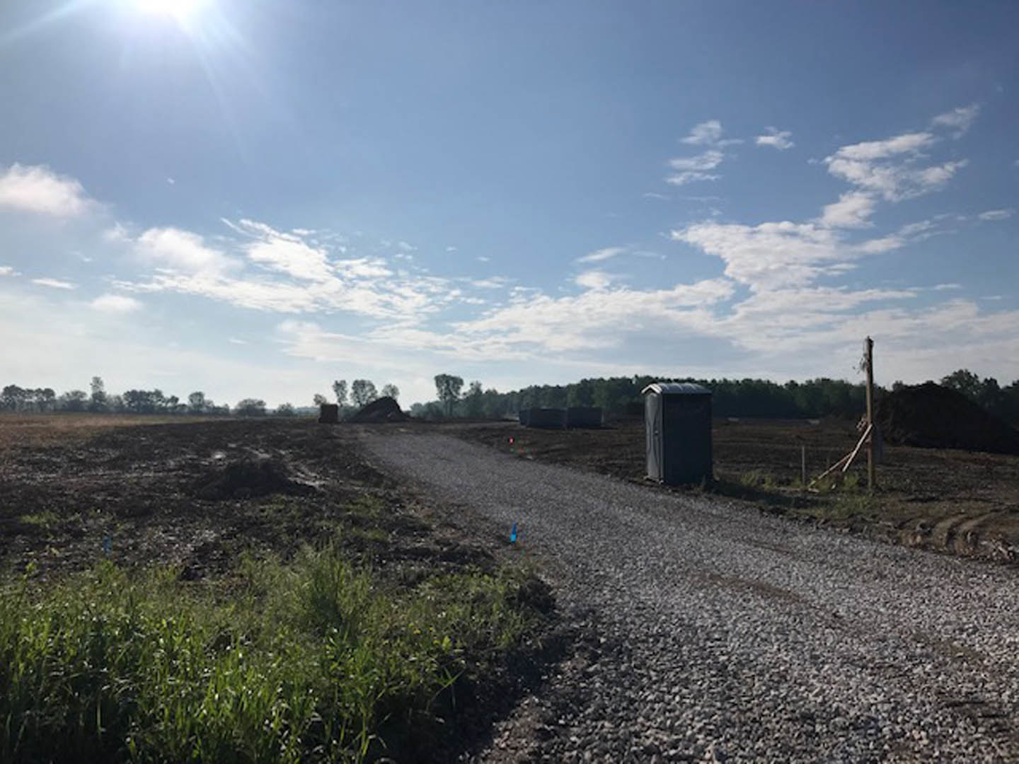 Gravel road bordered by tall grass leading to a small shed under a blue sky with scattered white clouds, sunlight highlighting the landscape.