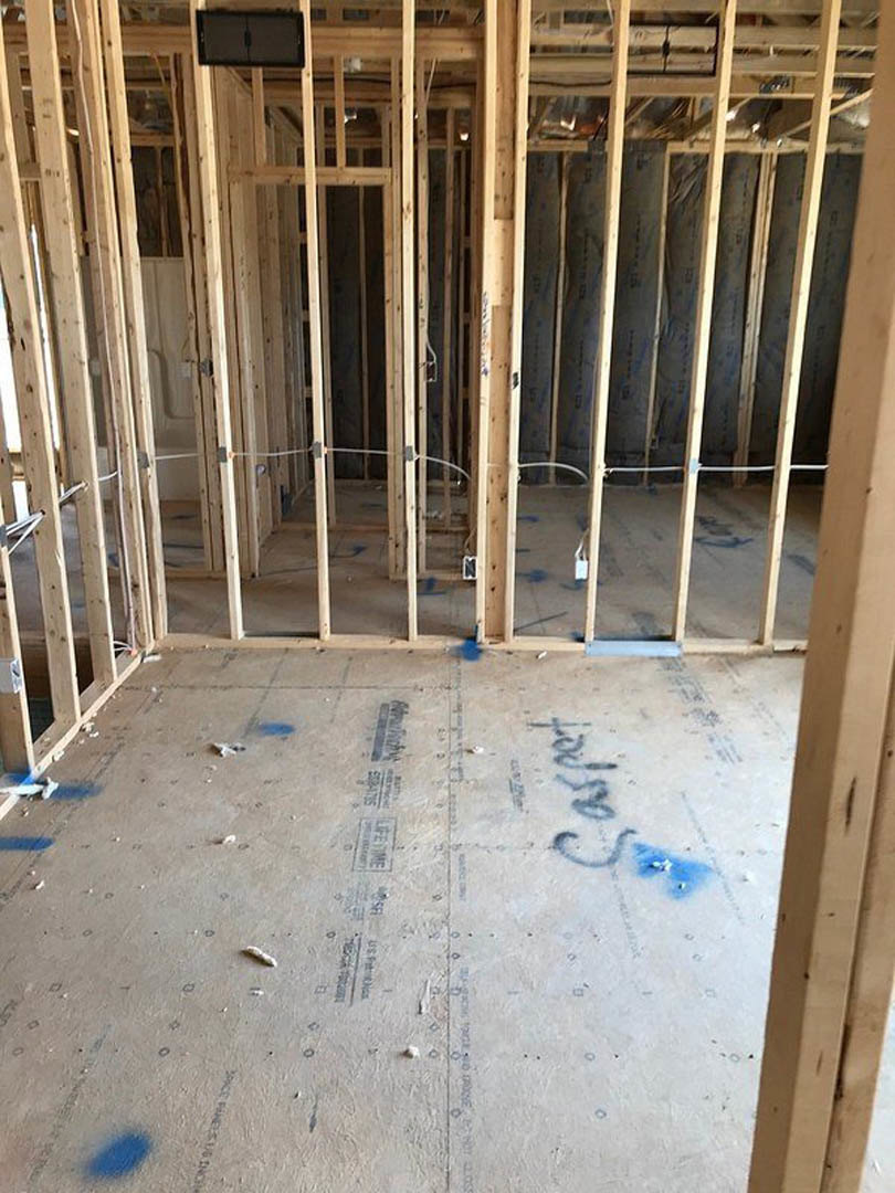Room under construction with exposed wood framing, concrete floor partially painted blue, black object resting on white surface, and construction materials scattered throughout.