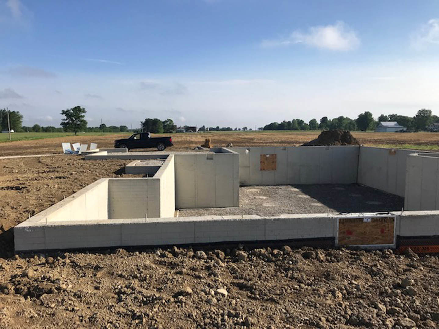 Dirt construction site with concrete foundation blocks, blue sky with scattered clouds, parked truck in background, and tree at edge of field