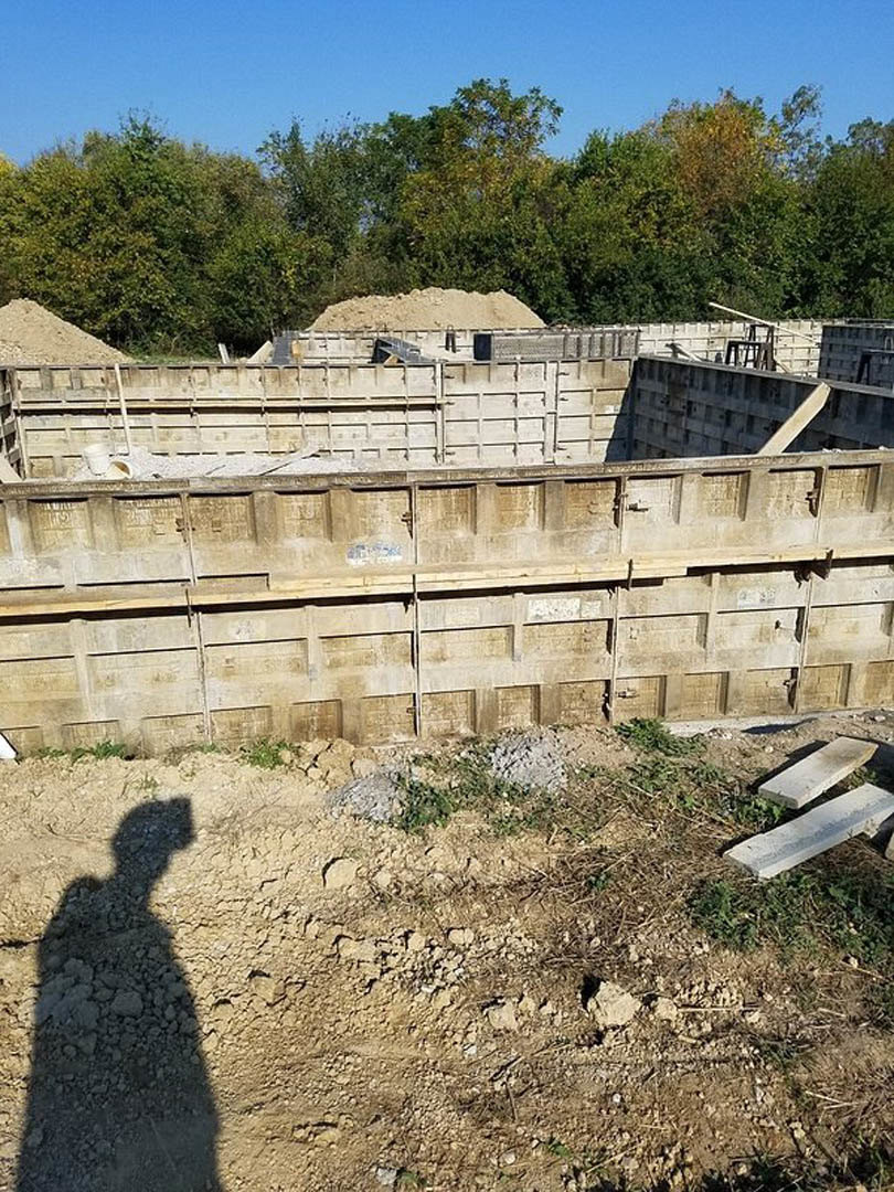 Concrete foundation under construction with exposed concrete walls, dirt surface showing a shadow, pile of sand near trees, white stone in foreground, blue sky overhead