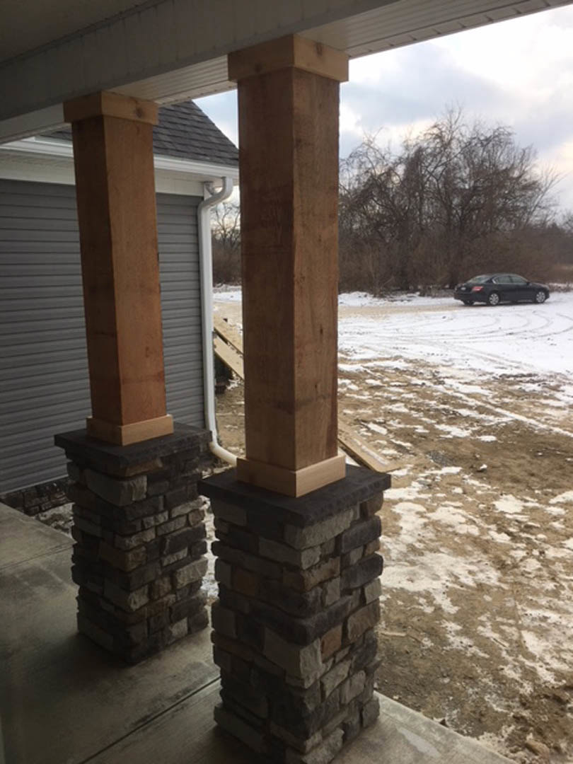 Stone porch pillars with brick bases, snow-covered ground, leafless trees, and a black car driving on the road in front of the custom home