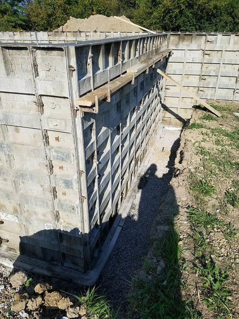 Concrete exterior wall casting a shadow onto adjacent ground, bordered by patches of grass and dirt, with metal bars and a wooden plank visible along the wall.