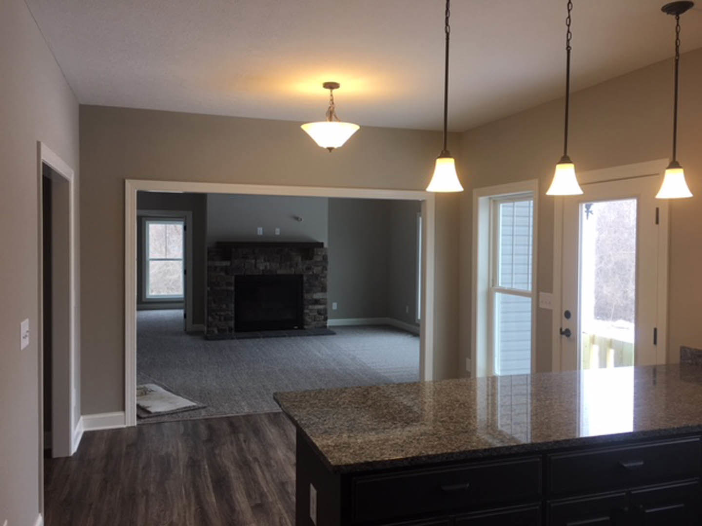 Open-concept kitchen and living room featuring stone fireplace, white cabinetry, wood flooring, pendant light fixture, and decorative pillows near a window with white tile trim