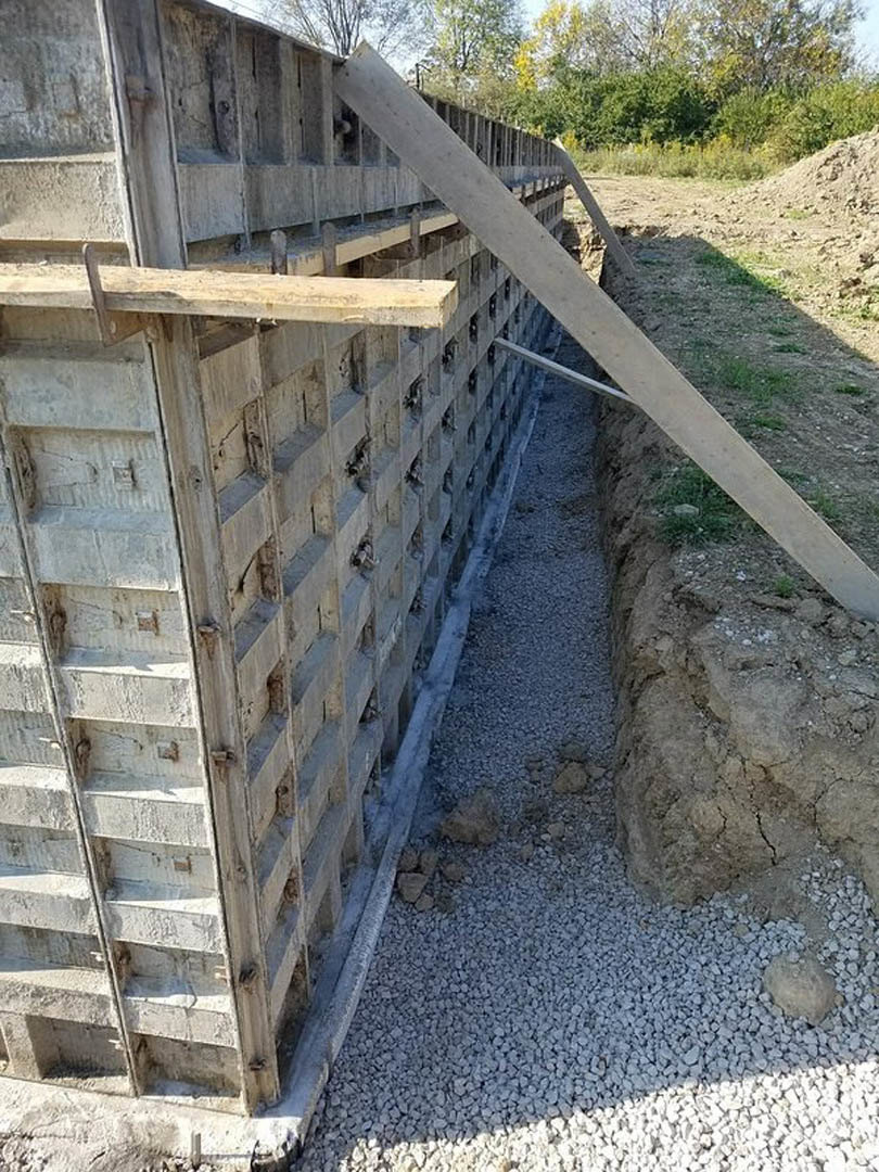 Concrete exterior wall with horizontal wooden beam, surrounded by rocks and gravel, adjacent to outdoor landscaping with plants and trees.