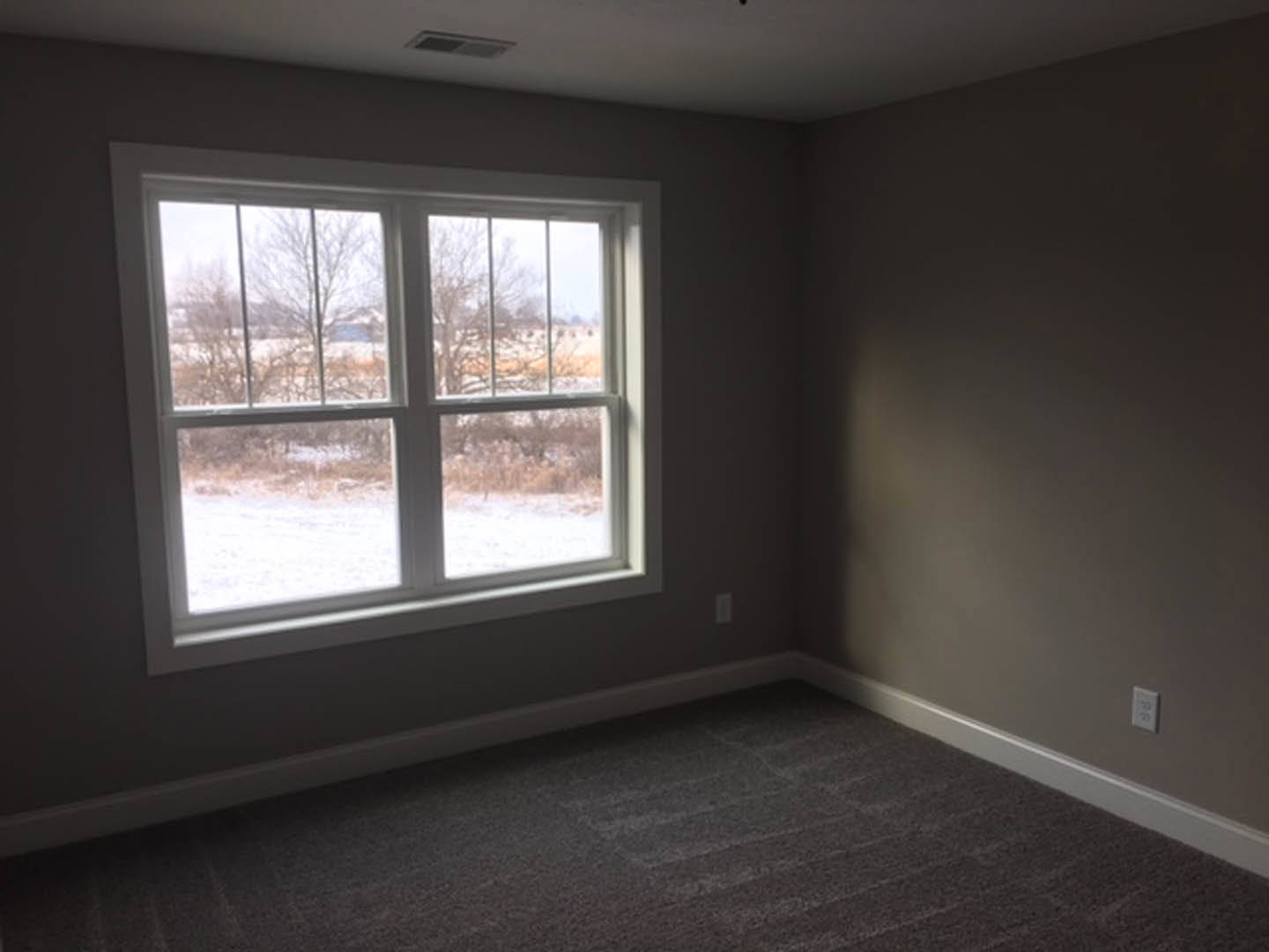 Carpeted room with large window overlooking snowy field, white electrical outlet with numbered label, floor vent, and trees visible outside