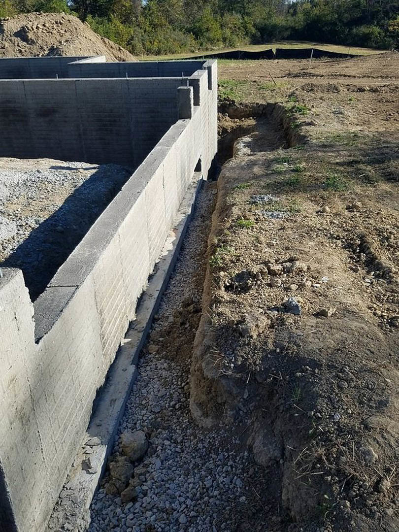 Concrete foundation wall with a drainage channel and curb, surrounded by gravel, dirt, and grass, with trees in the background.