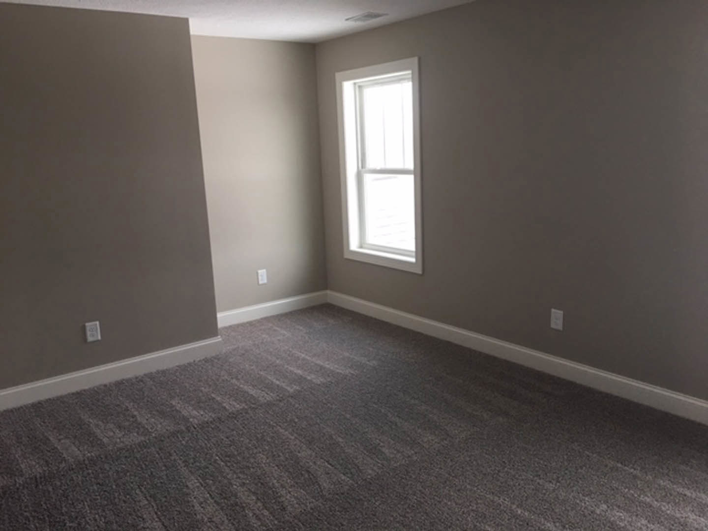 Carpeted room with white baseboards, large window letting in natural light, white electrical outlet on smooth wall, neutral finishes throughout