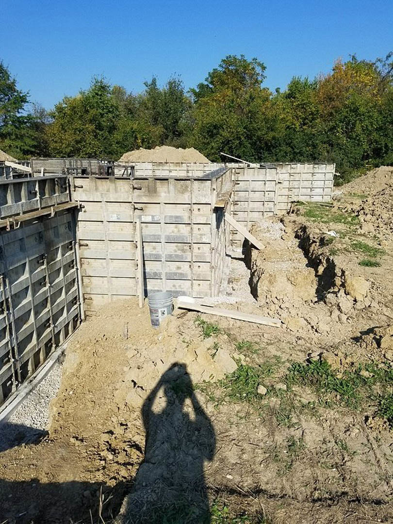 Shadow of a person cast on bare soil near a concrete wall, with a paint bucket on the ground, pile of sand, wooden plank, and trees in the background