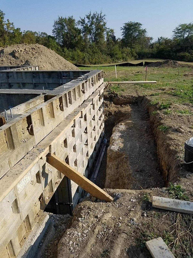 Concrete foundation walls with circular form holes, wooden beam propped against wall, grassy field and cluster of trees in background, soil and stone scattered around construction
