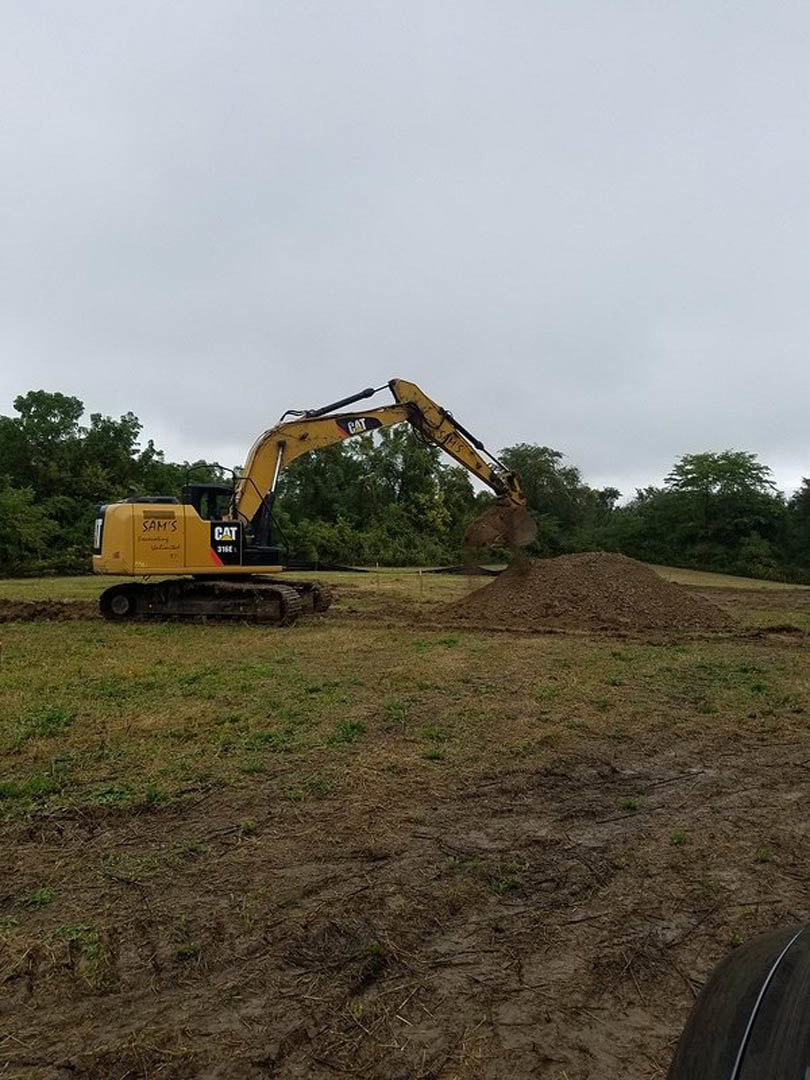 Yellow excavator with extended bucket digging into a dirt pile on grassy outdoor construction site, surrounded by trees and blue sky