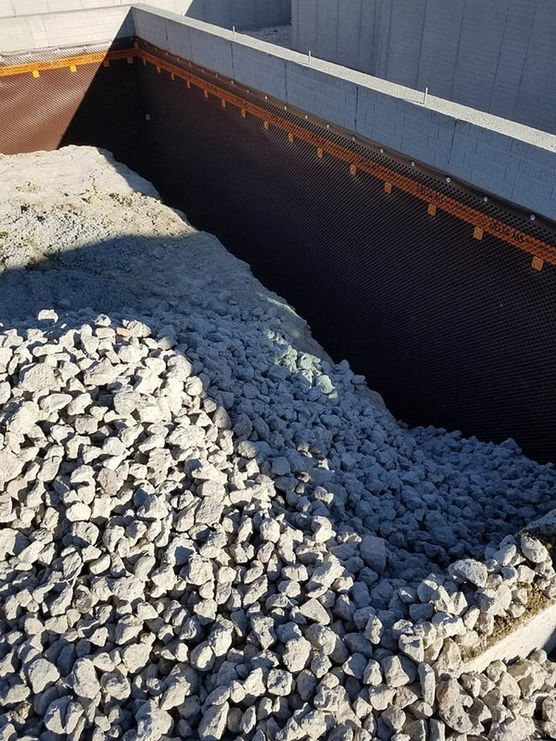 Gravel and large rocks stacked beside a composite exterior wall, with ground cover and outdoor landscaping visible