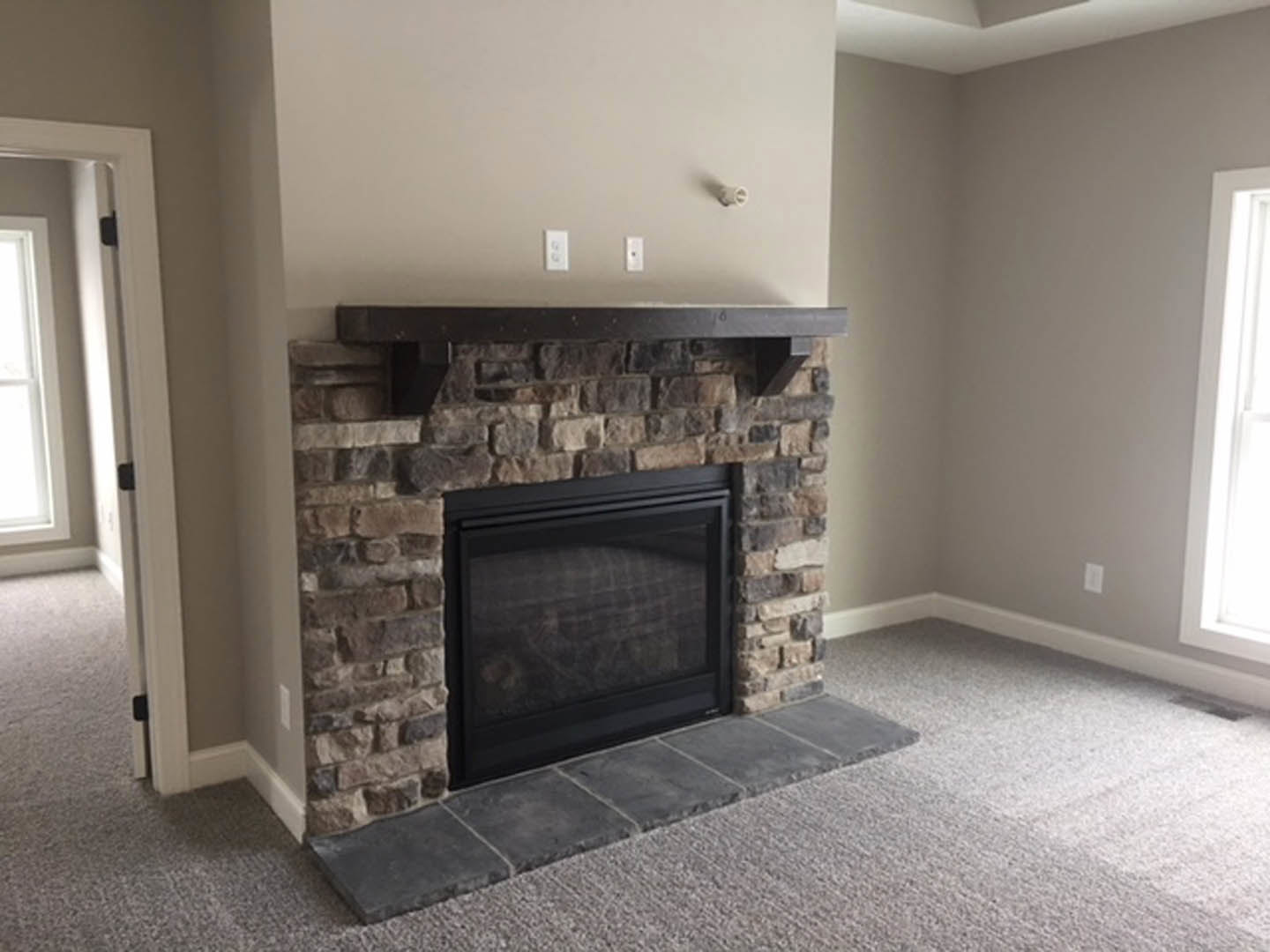 Stone hearth fireplace with glass door, wood mantel shelf, white wall, and window with white frame in a residential living space.