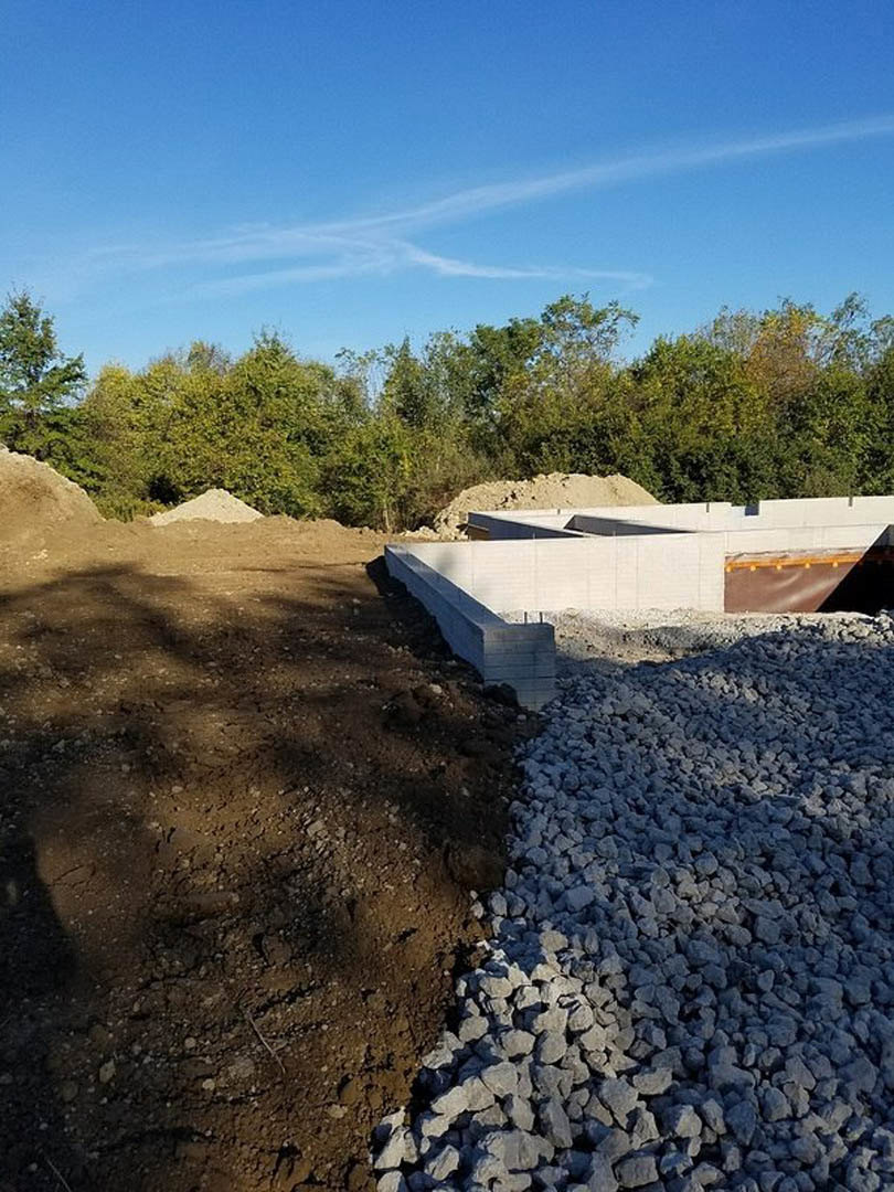 Gravel-covered construction site with a pile of rocks beside a brick wall, white wall featuring a hole in the ground, group of trees in the background, and blue sky with scattered