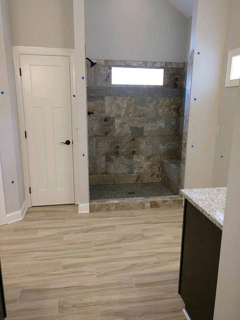 Bathroom with glass shower enclosure, wood plank flooring, stone accent wall with window, white door featuring black handle, white rectangular mirror with black border, white
