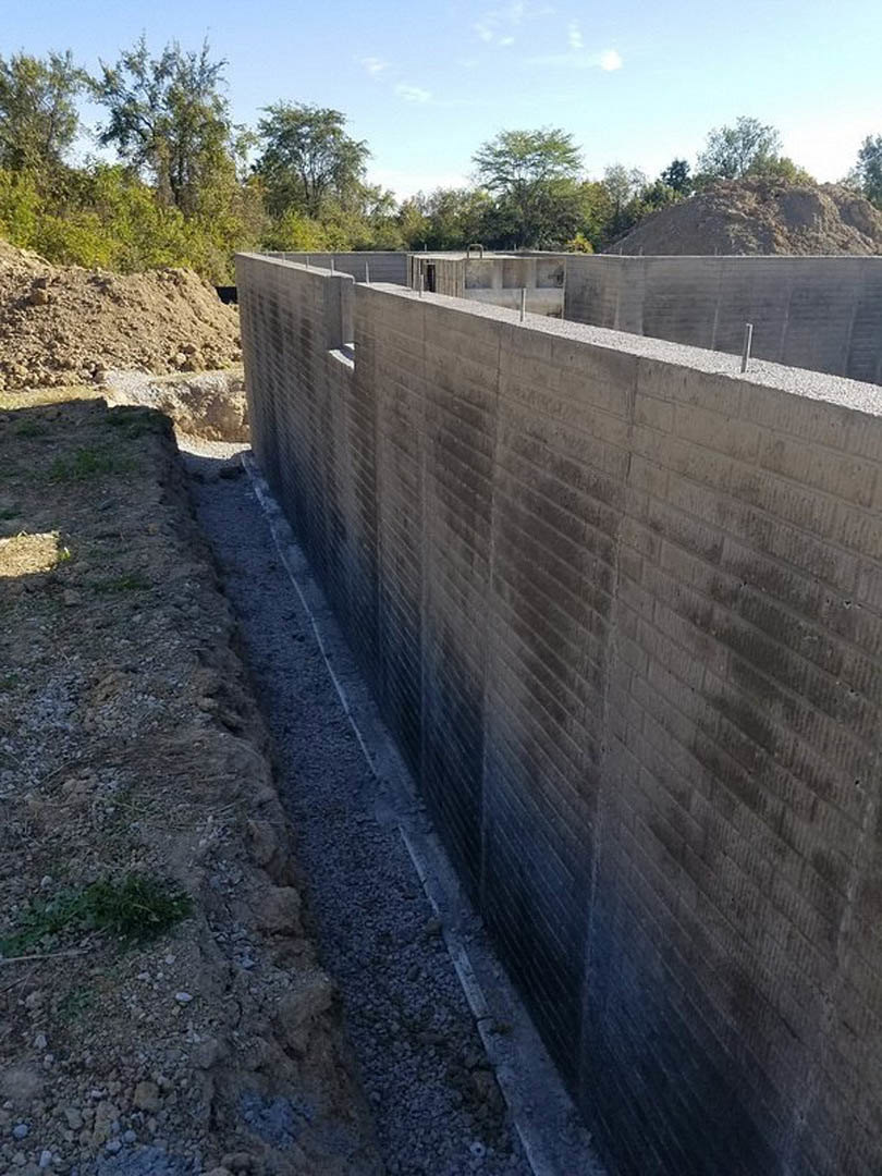 Concrete retaining wall with circular hole, exposed dirt pile in foreground, trees and blue sky in background
