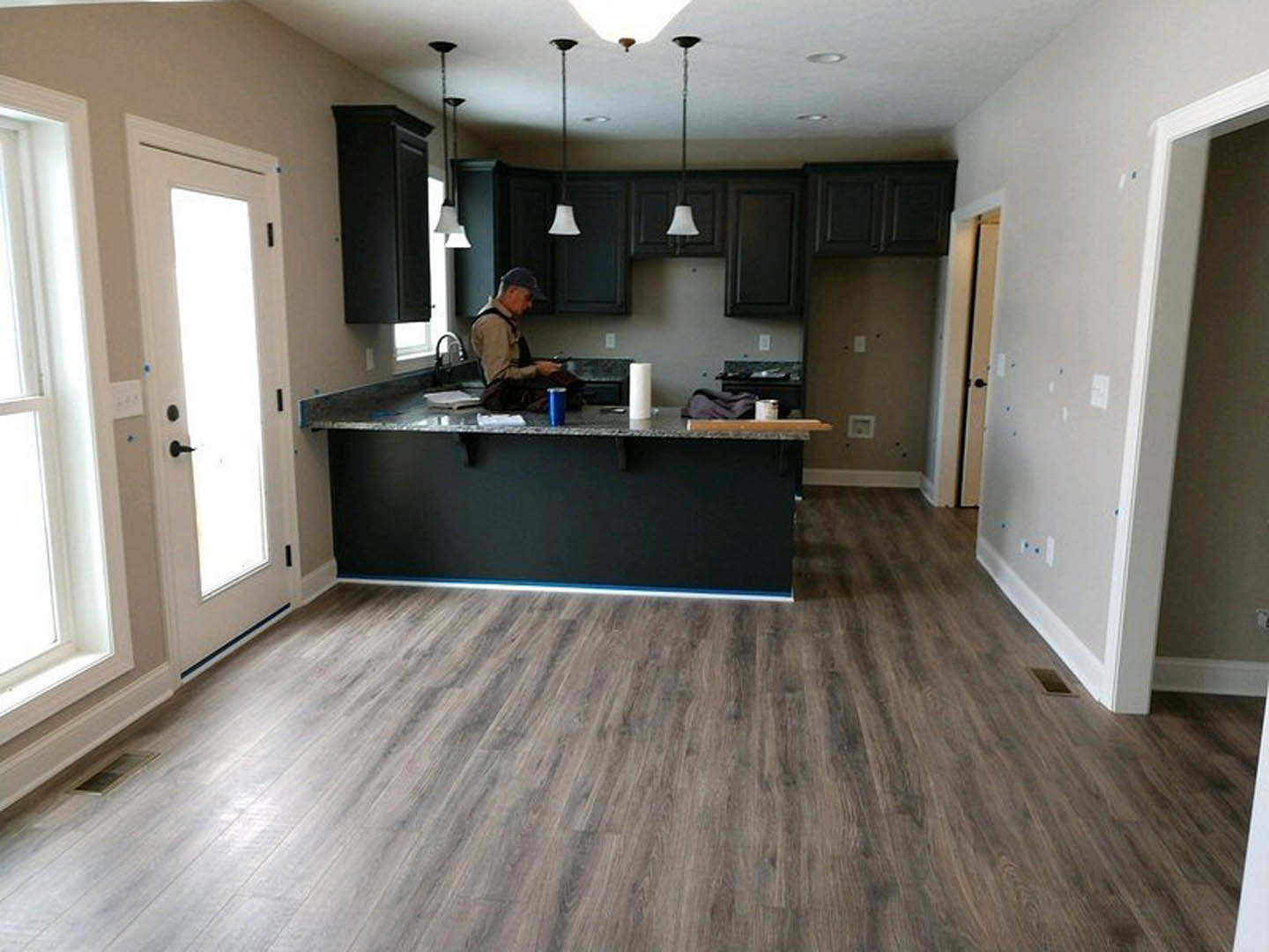 Man in hat and suspenders seated at kitchen counter with wood flooring, black accent wall, white cabinetry, and white door illuminated by natural light
