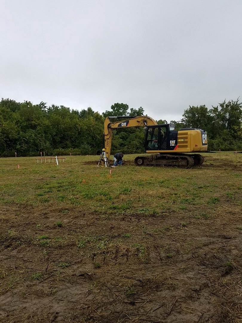 Yellow bulldozer with extended arm parked on grassy field, man in workwear standing nearby, cloudy sky and trees in background