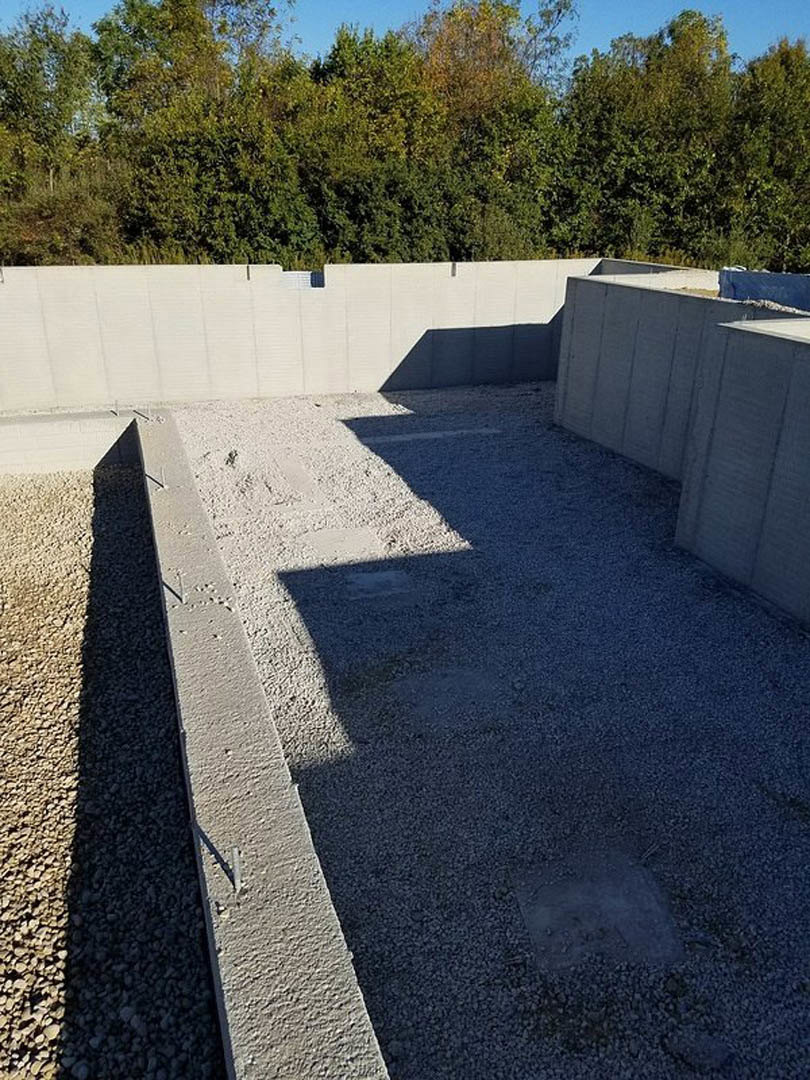 Concrete foundation bordered by gravel, surrounded by trees, with sunlight casting shadows on nearby white wall and rocks