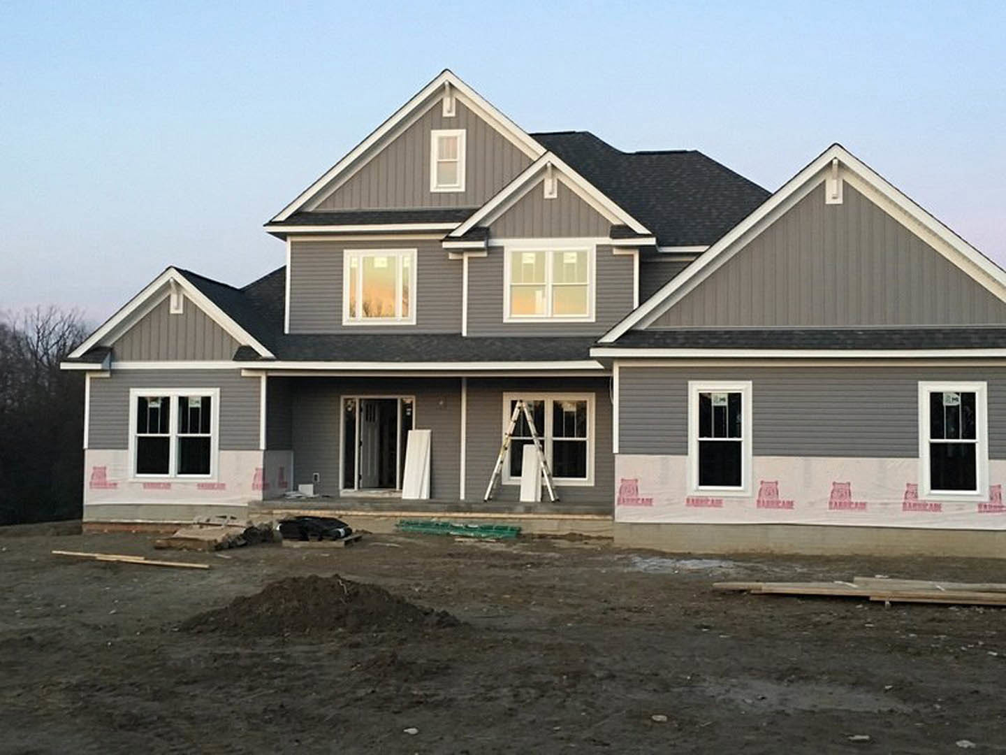 Two-story house under construction with exposed framing, white window frames, unfinished siding, dirt yard, and ladder leaning against exterior wall