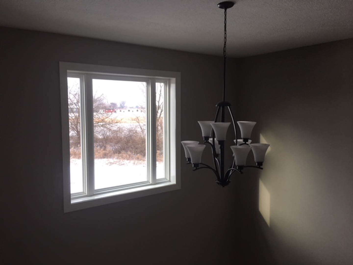 Three-light chandelier with white lampshades hanging from a plaster ceiling in a room featuring a large window with blinds and a view of a field and trees.