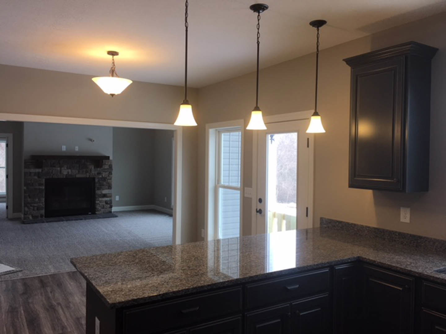 Open-concept kitchen featuring a stone fireplace, bar seating, television mounted above cabinetry, light fixtures on ceiling, wood drawers and doors, and polished countertops