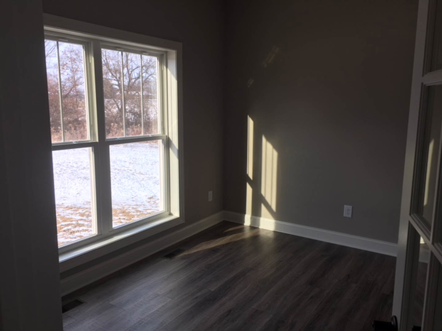 Sunlit room featuring a large window overlooking a snowy field, dark wood laminate flooring, and neutral gray walls.
